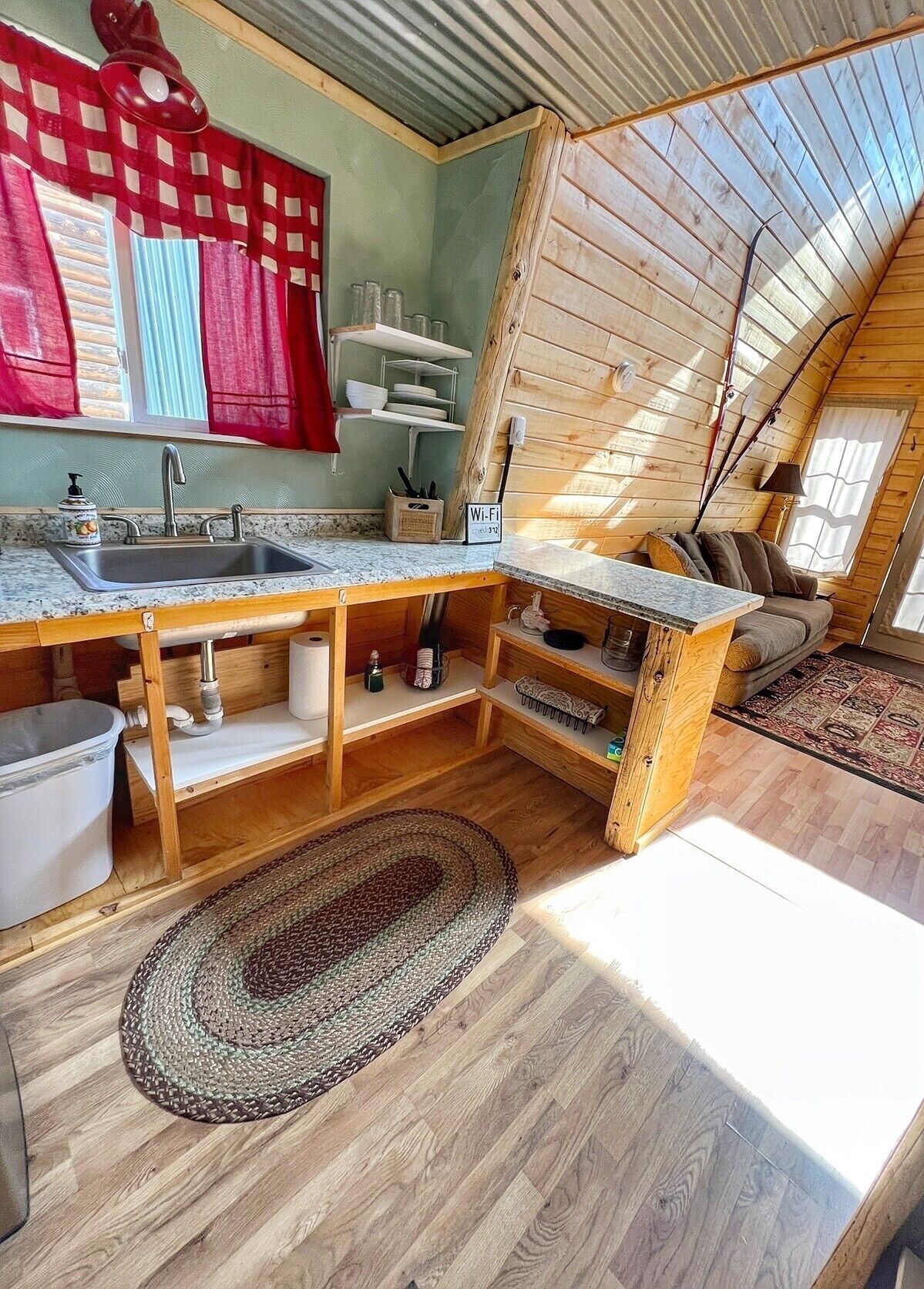 Una cucina compatta con lavello, piano di lavoro e pensili in legno. Si vede una finestra con tende rosse e un soffitto con travi in legno.