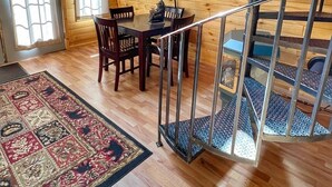 Ein Interieur einer Holzhütte mit einer Wendeltreppe, einem Essbereich inklusive Tisch und Stühlen sowie einem großen Fenster.