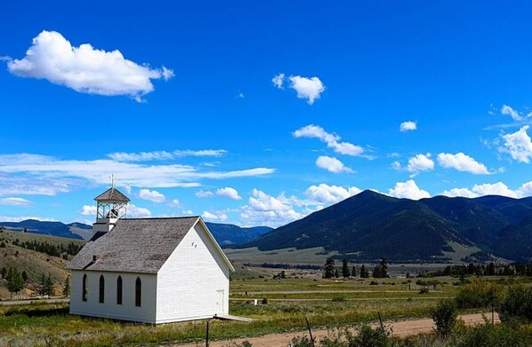 Une petite église blanche dotée d’un clocher, entourée d’un paysage rural avec des montagnes en arrière-plan.
