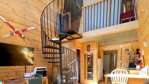 Interior de una cabaña de madera con una escalera en espiral, una zona de comedor, un sofá y un televisor.