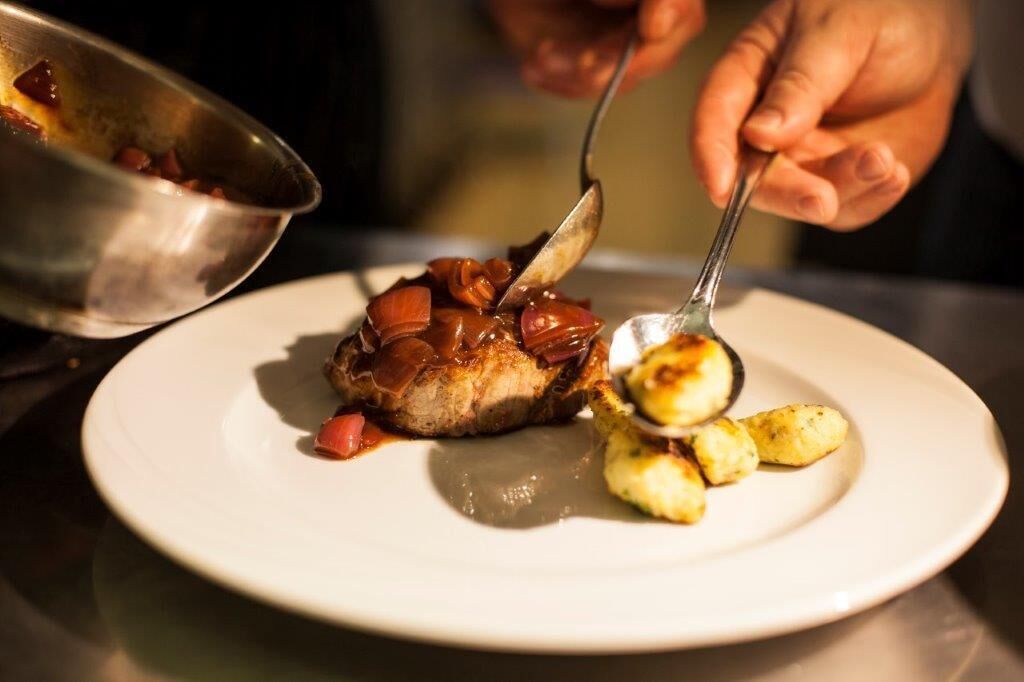 A person serving food onto a plate, including a piece of meat with a sauce and some vegetables.