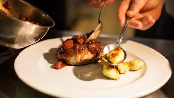 A person serving food onto a plate, including a piece of meat with a sauce and some vegetables.