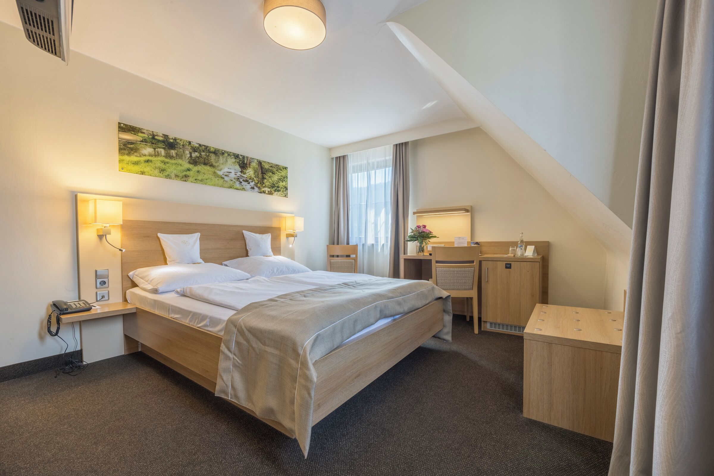 Ein Hotelzimmer mit Bett, Nachttischlampen, einem Schreibtisch mit Computer, Telefon und einem Bild an der Wand.