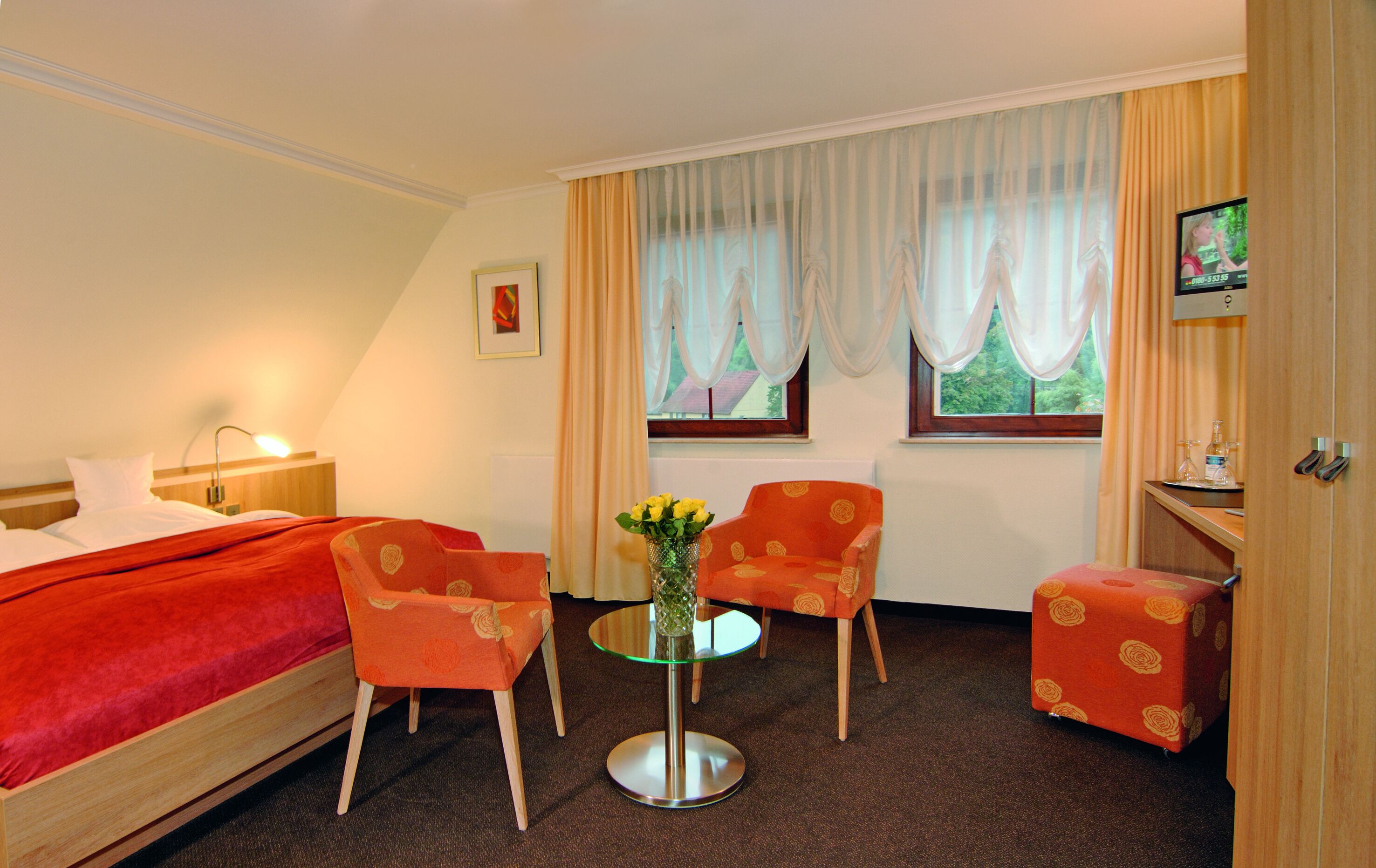 A hotel room with a bed, two armchairs, a small table with a vase of flowers, a TV, and a window with curtains.