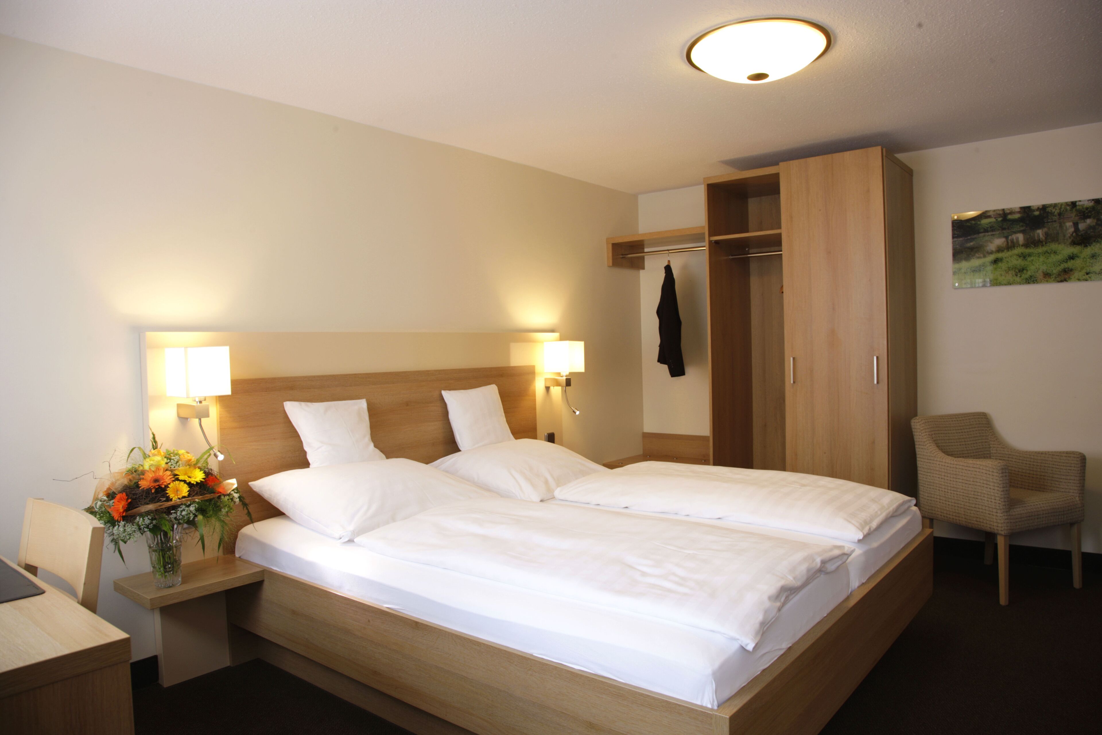 Ein Doppelbett mit zwei Kissen, ein Nachttisch mit Lampe, ein Sessel, ein Kleiderschrank und ein an der Wand hängendes Bild.