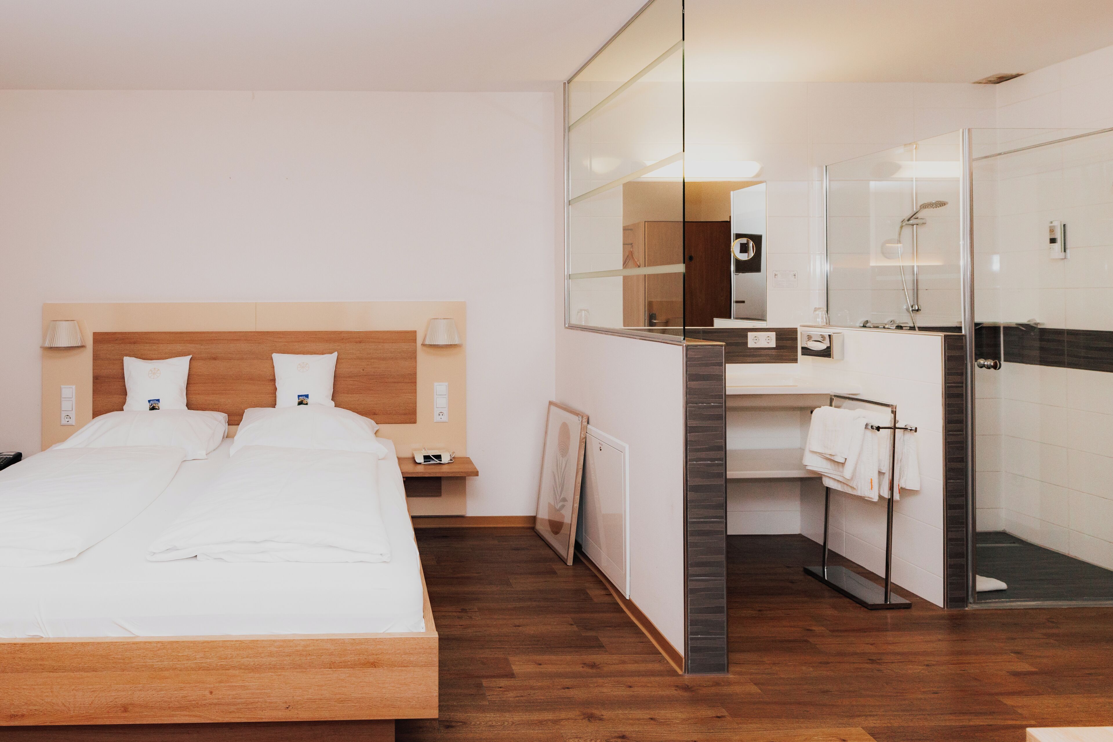 Una habitación de hotel con una cama de madera, un espejo grande, un baño con ducha y un perchero para toallas.