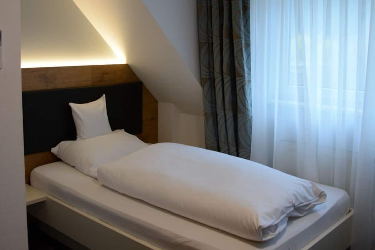 A modern hotel room with a bed, a headboard, a ceiling light, and a window with curtains.