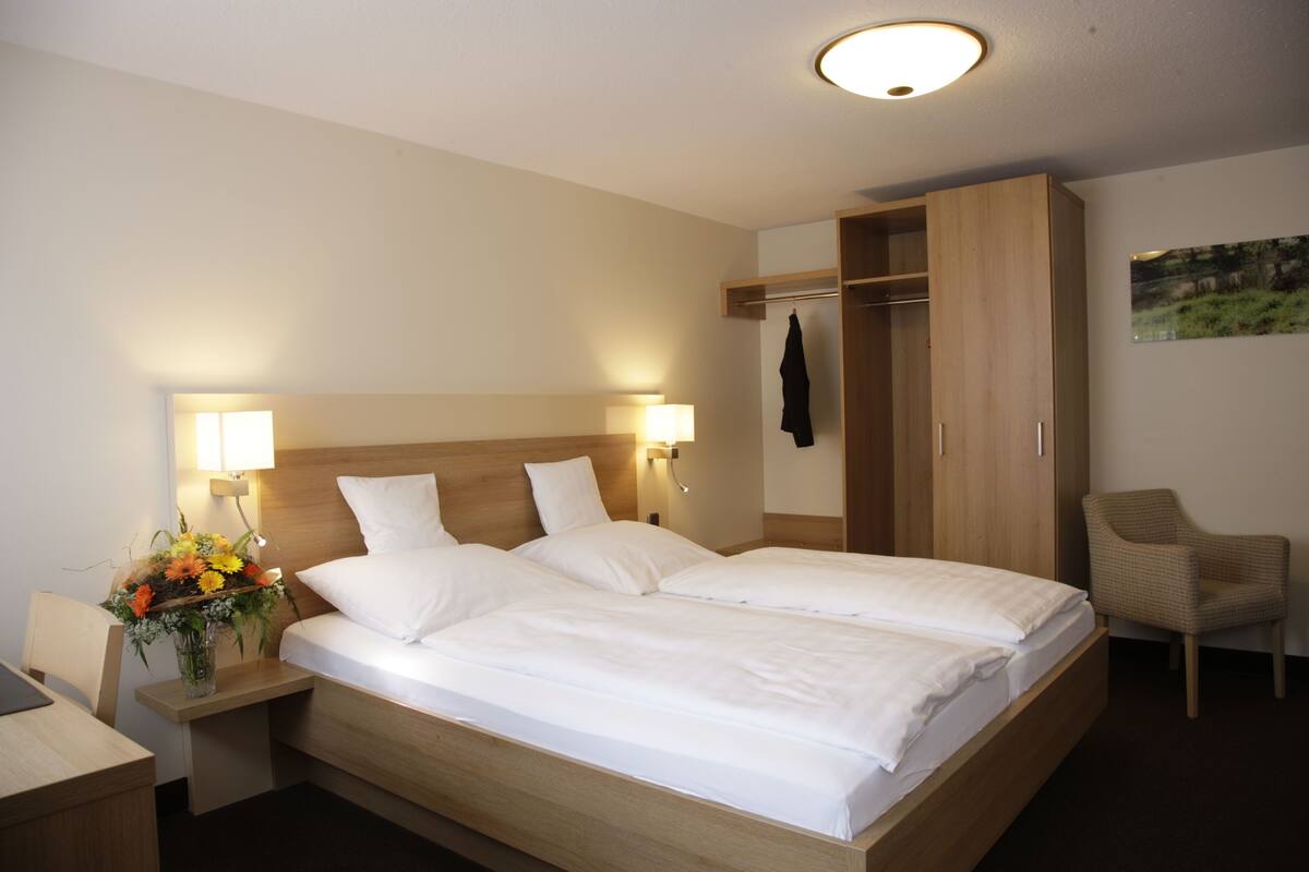 A hotel room with a double bed, bedside lamps, a wooden wardrobe, a chair, and a desk with a vase of flowers.