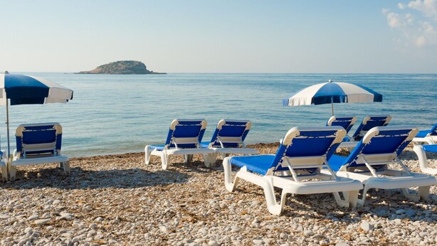 A beach with several sun loungers and umbrellas, a pebbled shore, and a distant island. - Lovely apartment WiFi, A/A, Parking, bike station, terrace & pool (Altea)