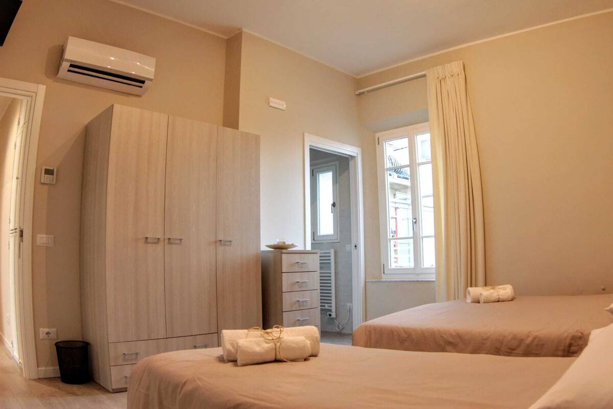 A hotel room with two beds, a wardrobe, an air conditioning unit, a radiator, and a window with curtains.