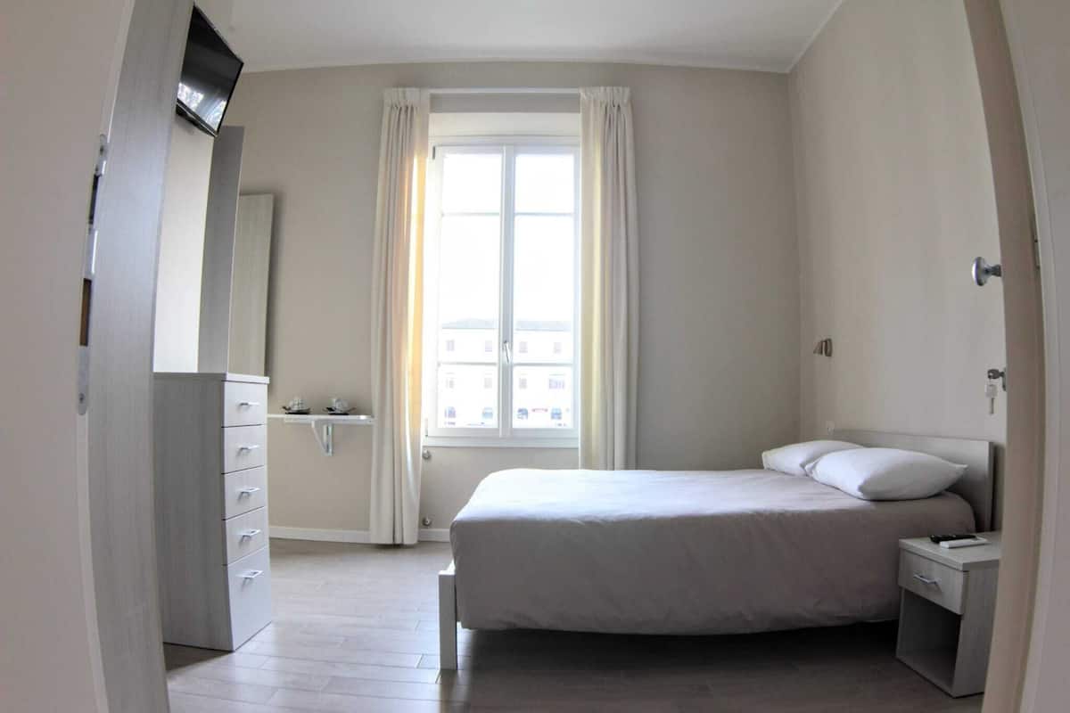 A bedroom with a bed, a nightstand, a dresser, a window with curtains, and a view of buildings outside.