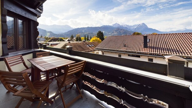 A balcony with wooden furniture, a view of a mountain range, and traditional architecture. - Fantastic central Zugspitze view with parking lot  (Garmisch-Partenkirchen)