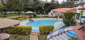 Une piscine entourée de haies bien entretenues et un espace patio avec des sièges d’extérieur.