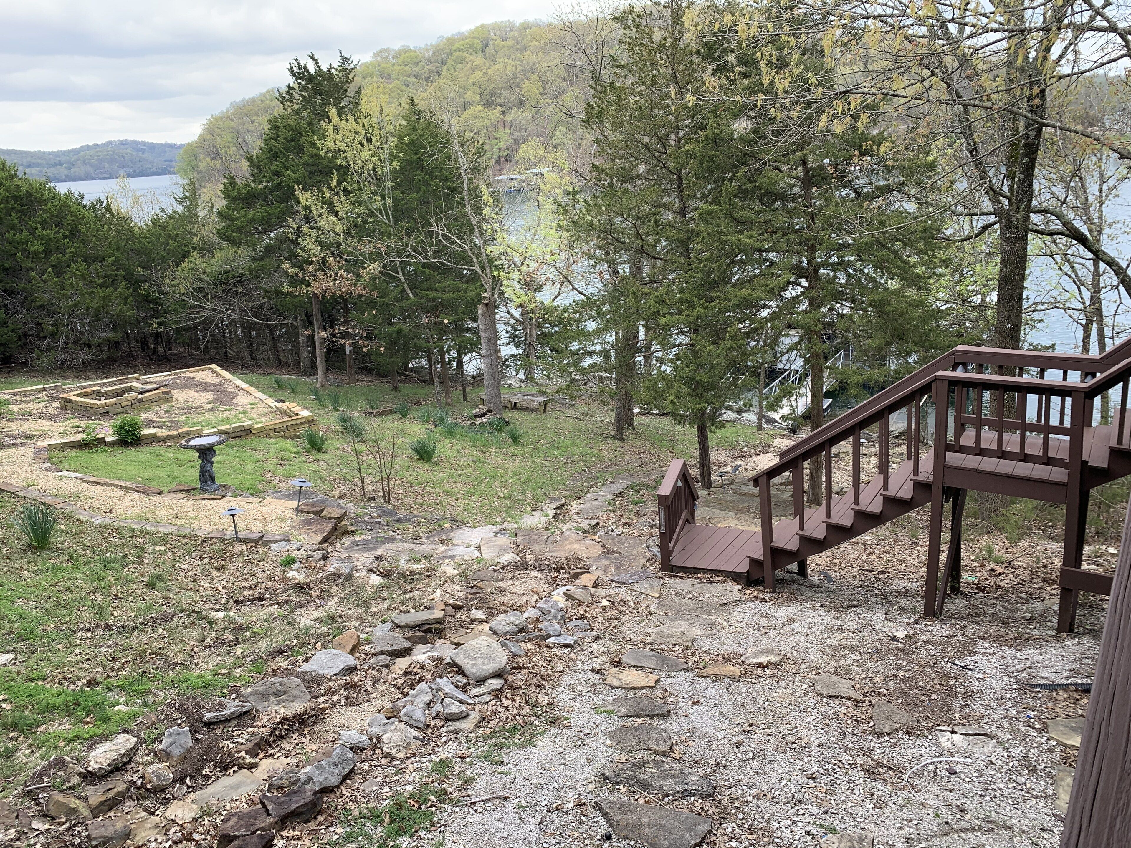 A wooden deck leading to a stone pathway surrounded by a forested area with a view of a lake.