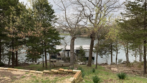A lakeside property with a stone deck, grassy areas, and trees.