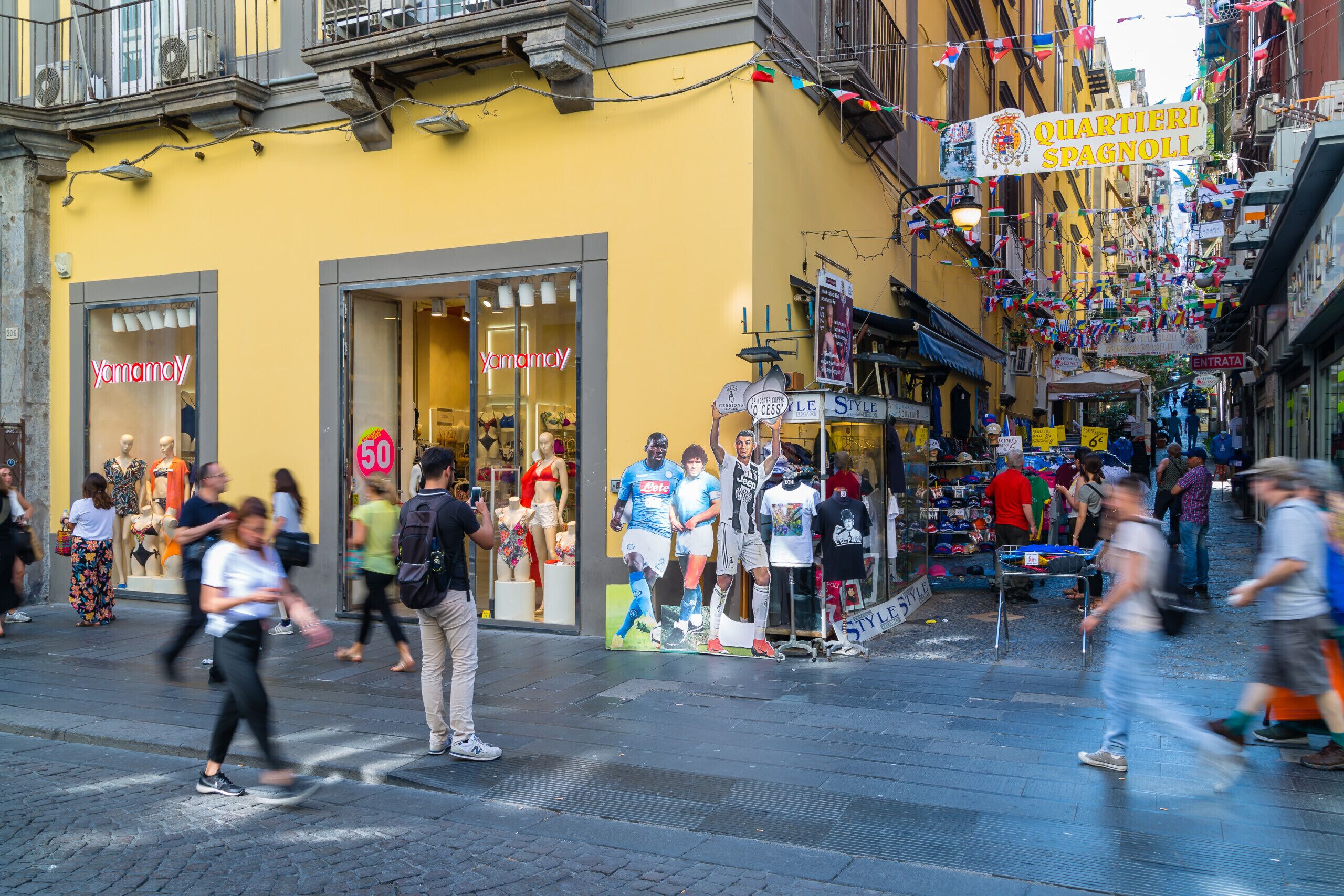Une rue animée, avec un bâtiment jaune abritant un magasin « Yamamay » et une enseigne indiquant « Quartieri Spagnoli ».