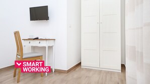 Una habitación con un escritorio blanco, una silla, un televisor montado en la pared y un armario blanco.