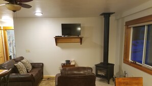 A living room with a sofa, a chair, a wooden table, a TV on a shelf, and a wood-burning stove.