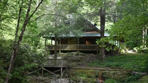 Eine Holzhütte mit einer Holzterrasse und einer Stegbrücke über einem Bach.