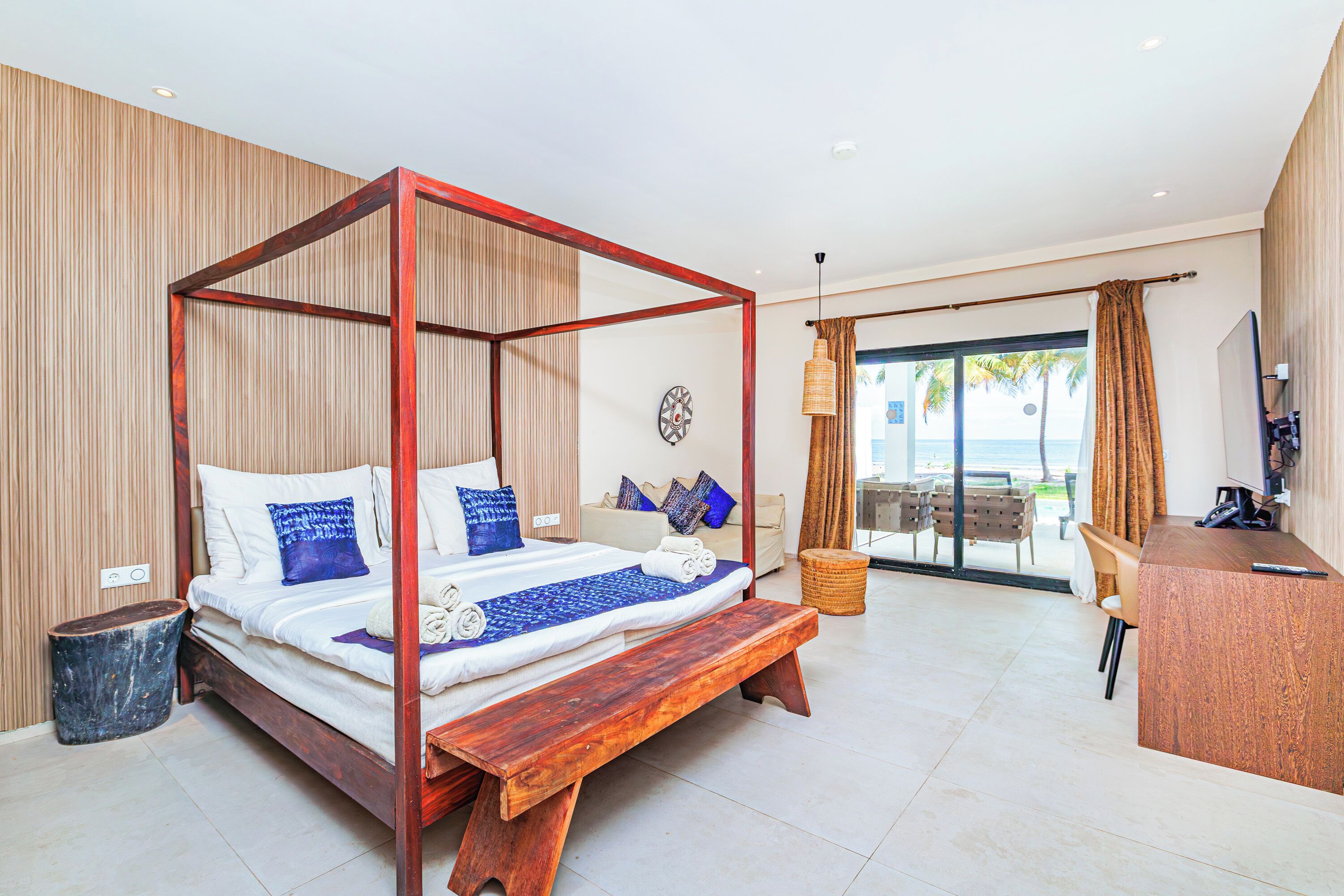 Een ruime slaapkamer met een hemelbed, een houten bankje, een bureau en uitzicht op het strand door de glazen deur.
