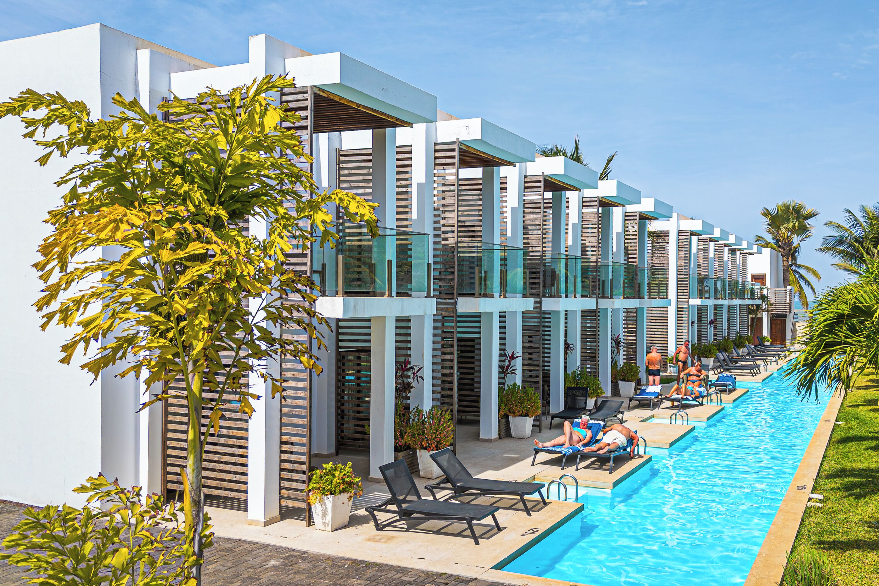Een modern hotel met een zwembad, ligbedden en een helderblauwe lucht.