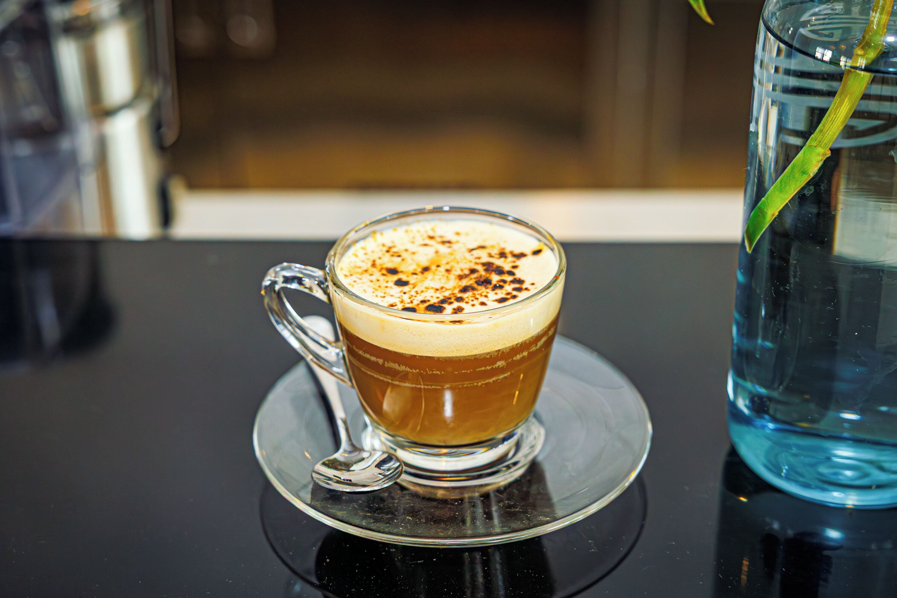 A clear glass cup of coffee with a saucer on a reflective surface.