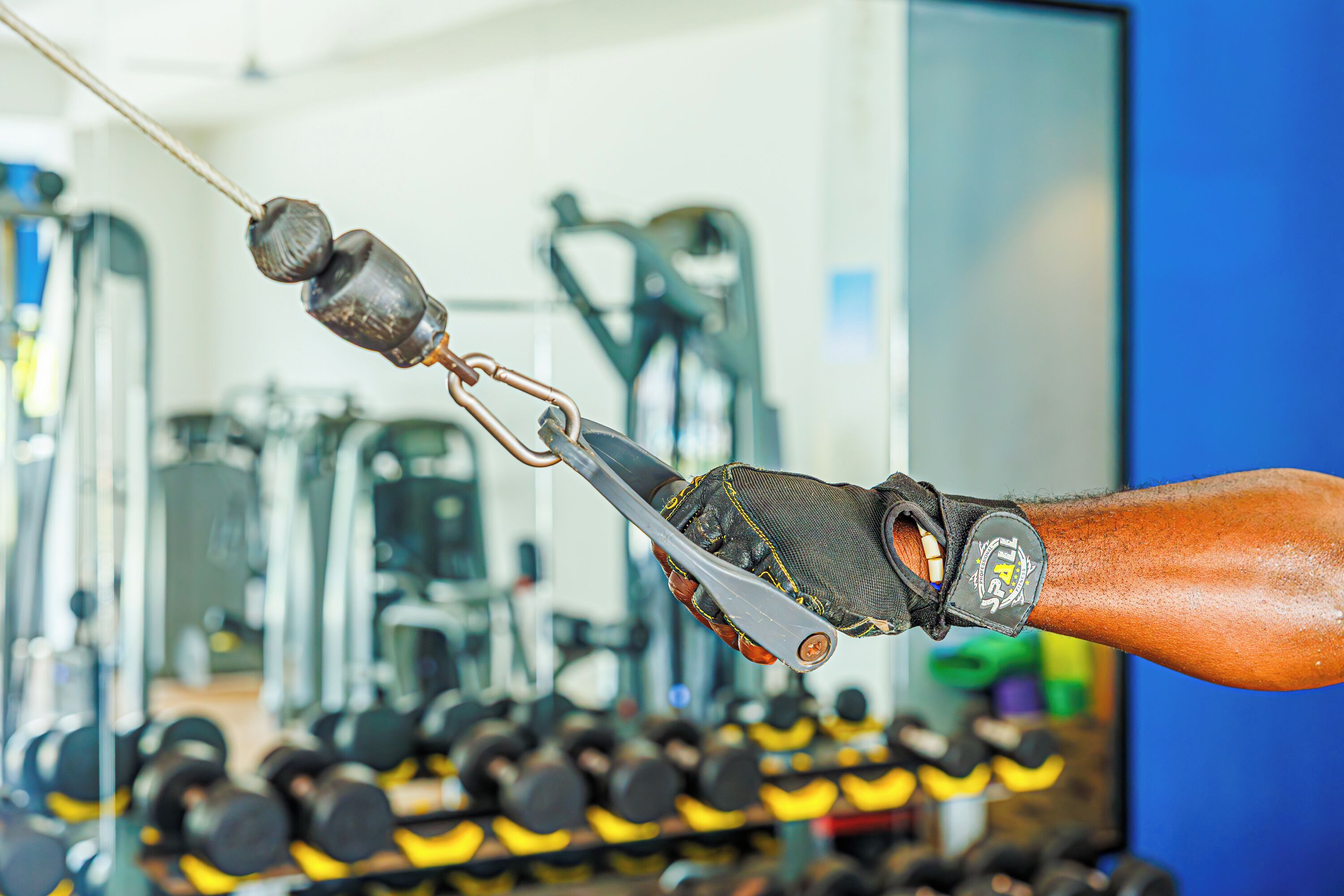 A close-up of a hand gripping a weightlifting cable with a weight attached, in a gym setting with exercise equipment and dumbbells in the background.