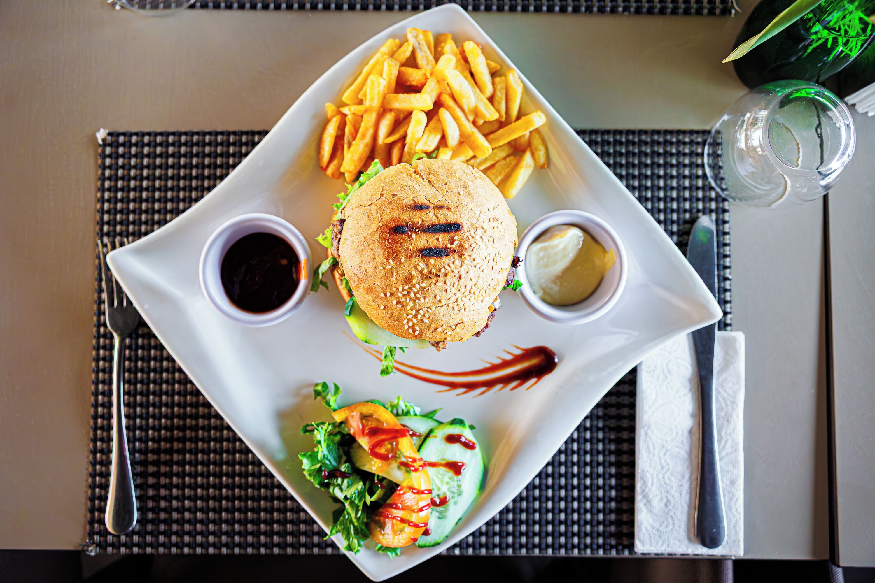 A square plate with a burger, fries, and a salad, accompanied by two sauces and a napkin.