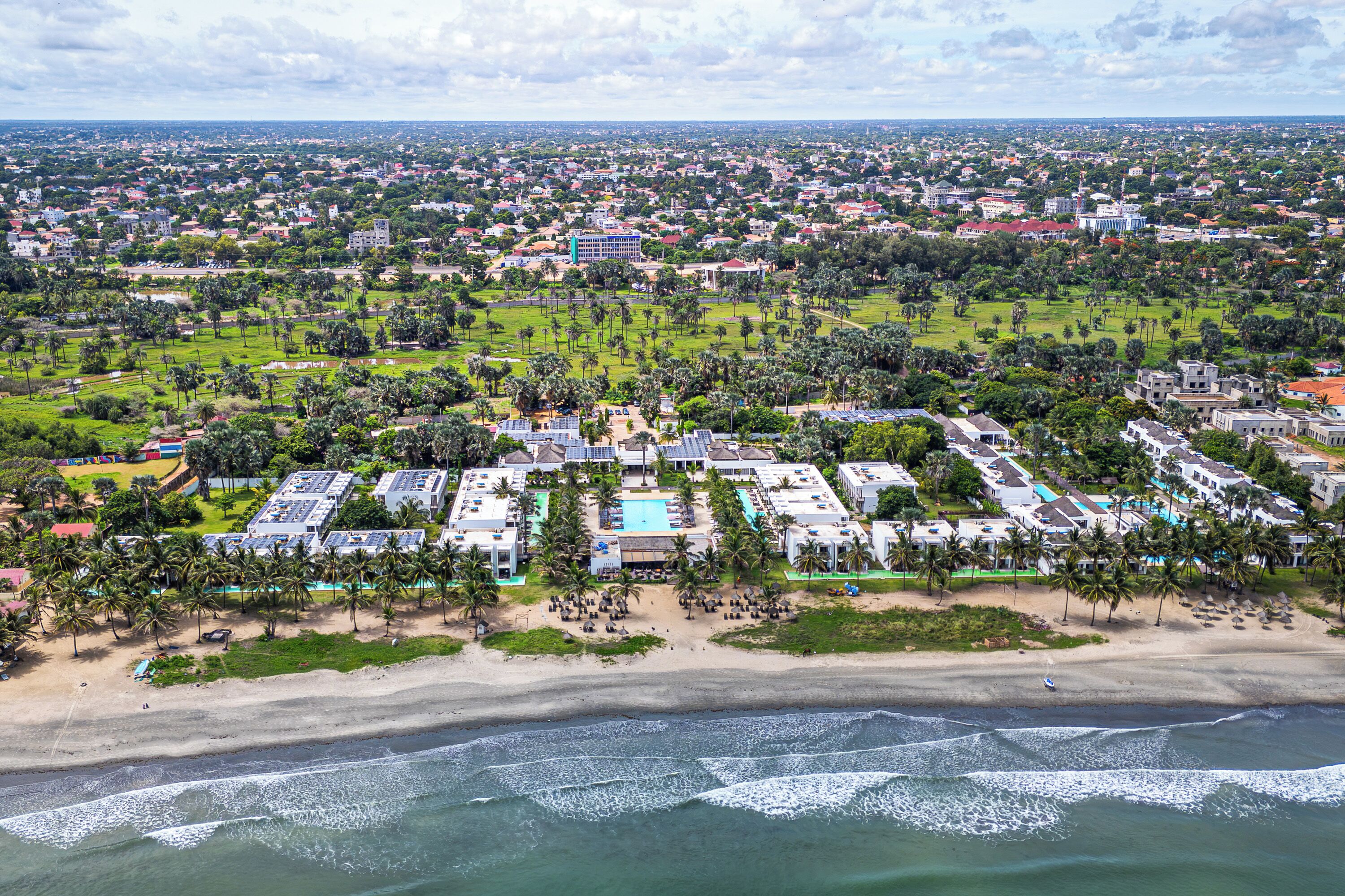 Un complejo turístico frente al mar con varias construcciones, una playa y vista a la ciudad de fondo.
