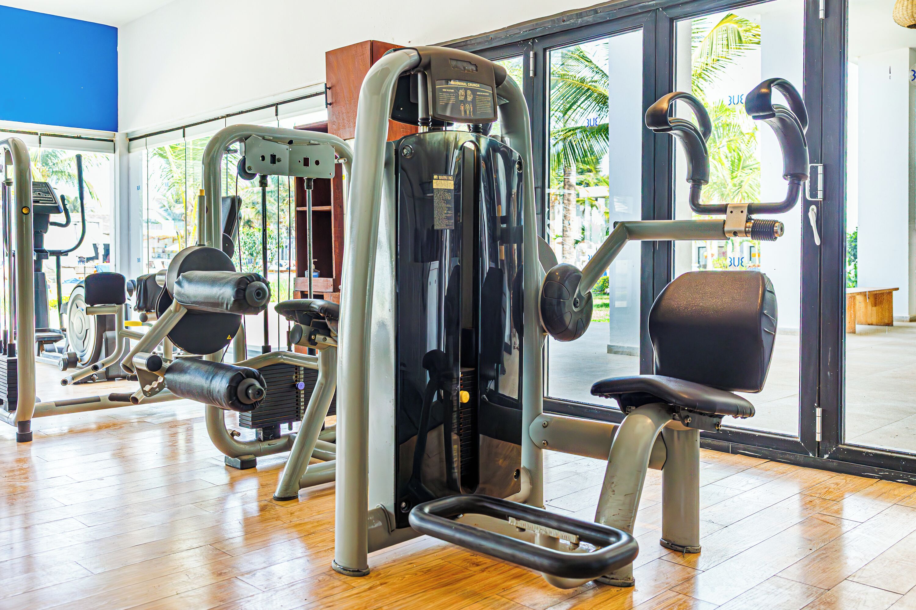 A gym with various exercise machines, a wooden floor, and large windows with a view of palm trees.