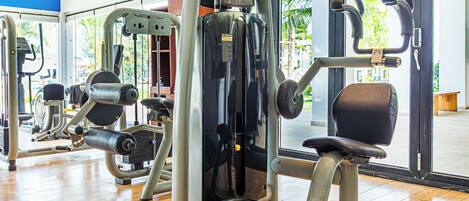 A gym with various exercise machines, a wooden floor, and large windows with a view of palm trees.