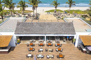 Un restaurante frente al mar con terraza al aire libre, vista al océano, palmeras y una playa de arena.