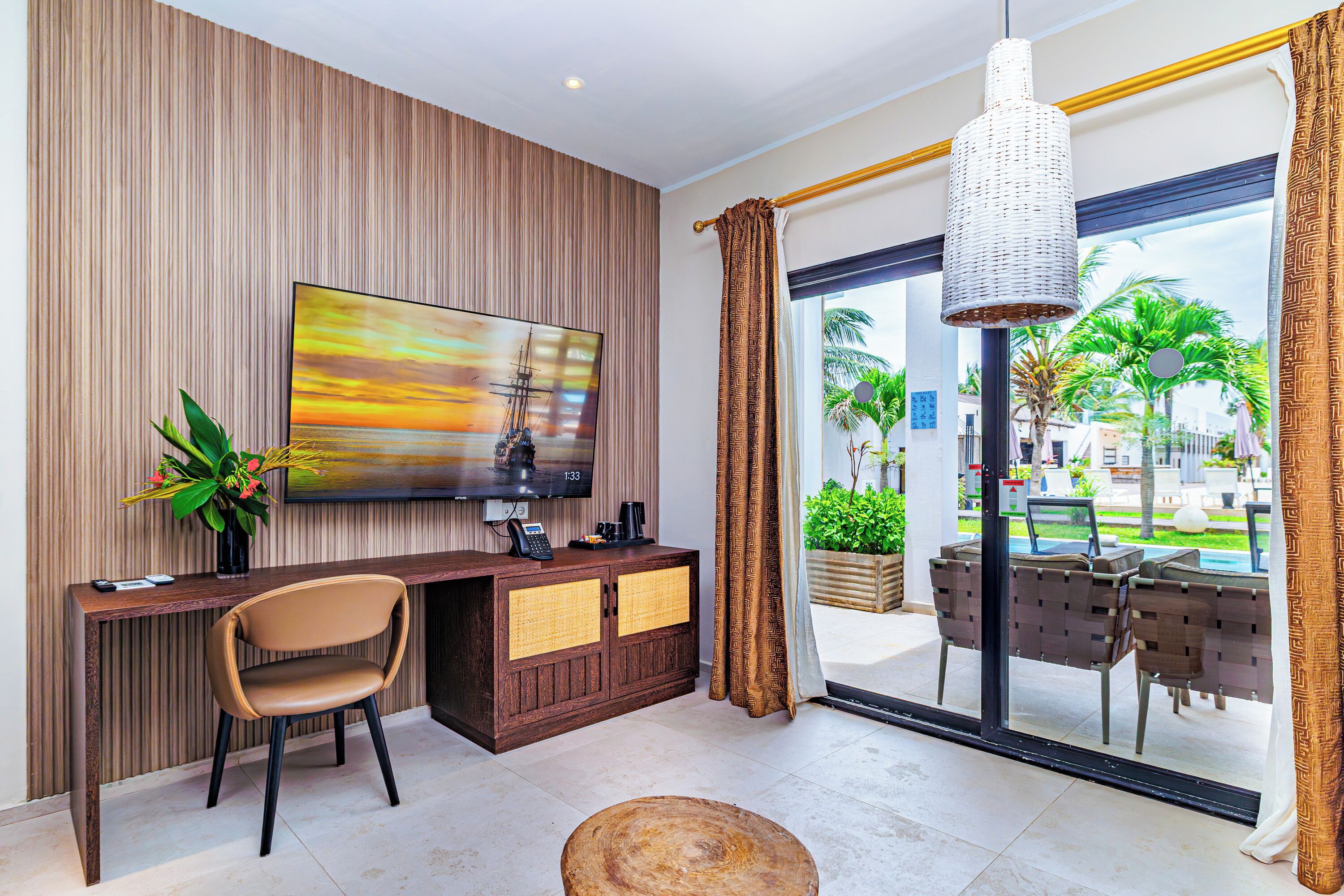 A modern hotel room with a large TV, wooden panel wall, a desk with a chair, and a view of palm trees through the glass door.