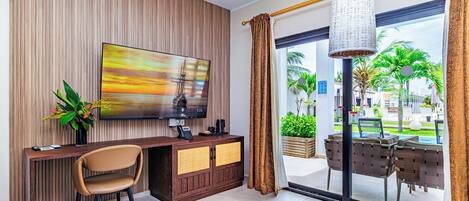 A modern hotel room with a large TV, wooden panel wall, a desk with a chair, and a view of palm trees through the glass door.