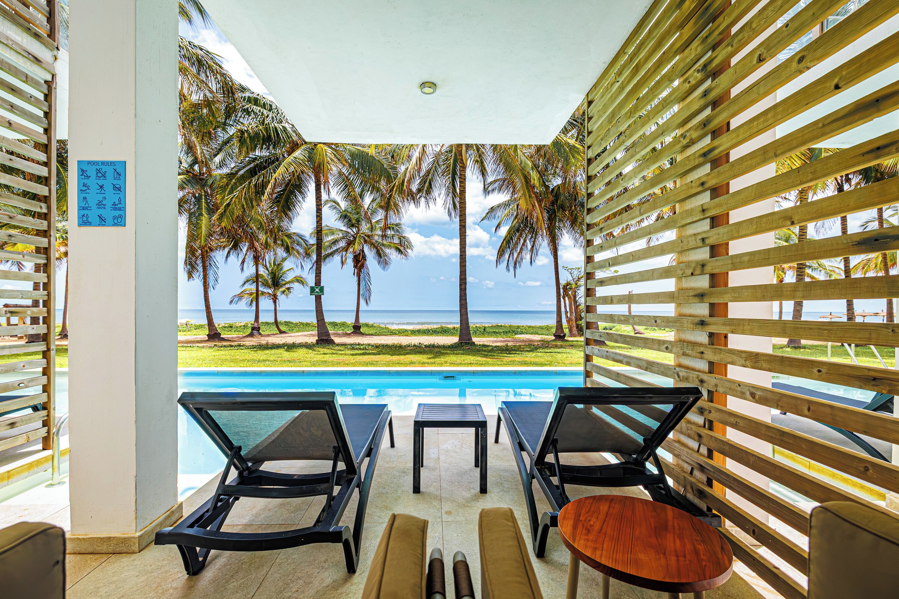Un espace piscine avec des chaises longues et une table, offrant une vue sur une plage bordée de palmiers et sur l’océan.