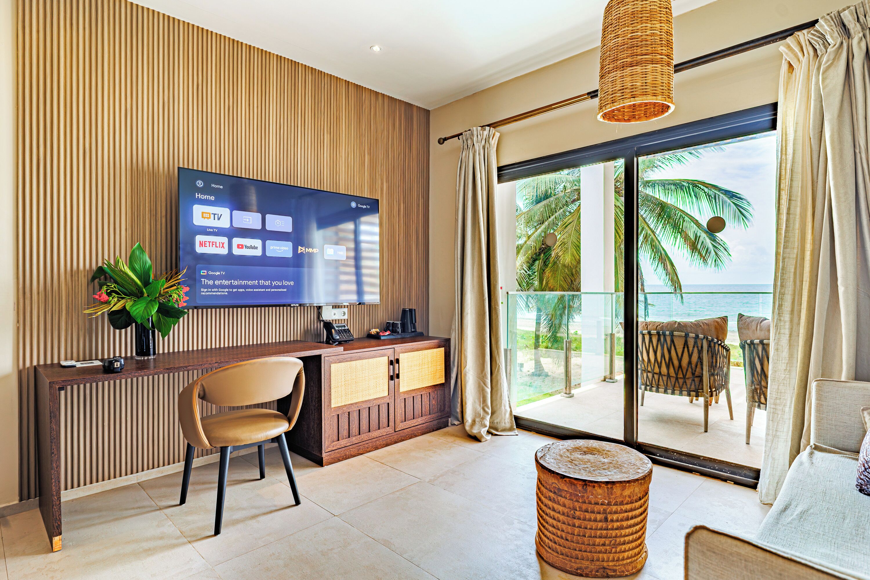 A modern hotel room with a flat-screen TV displaying streaming services, a wooden console table, a seating area with a view of the ocean, and a balcony with outdoor furniture.