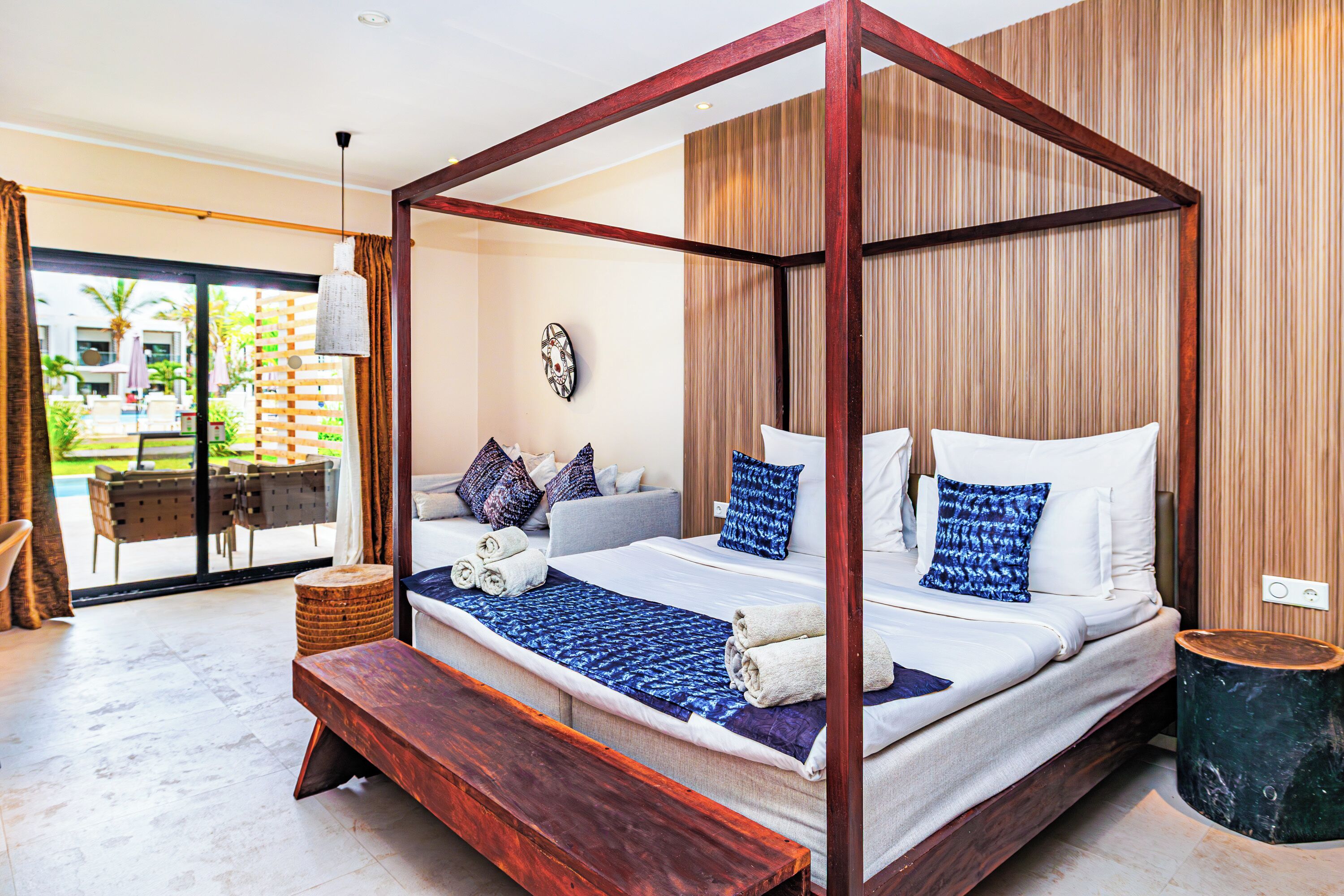 A four-poster bed with a wooden frame, a bench, and a small round table in a modern room with a large window and a view of a pool and greenery outside.