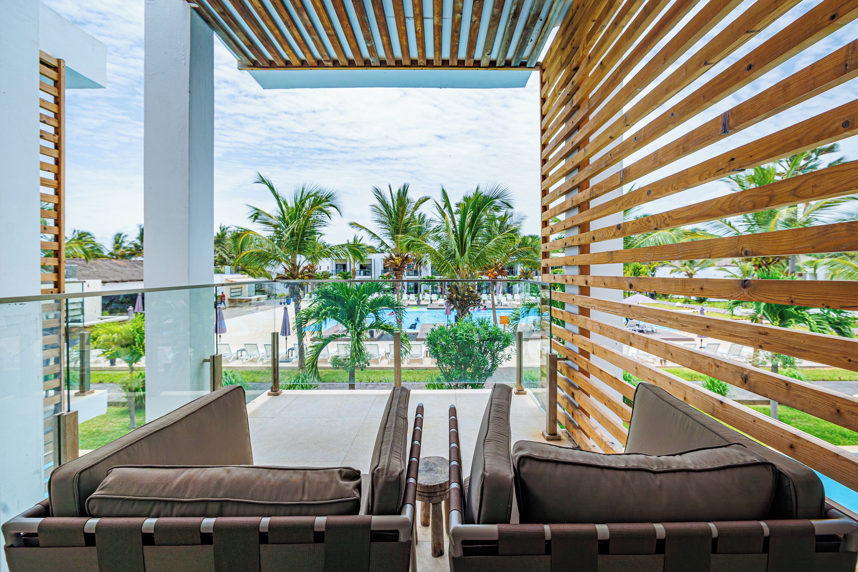 Un balcón con listones de madera, dos sillas acolchadas y vistas a una zona de piscina con palmeras.