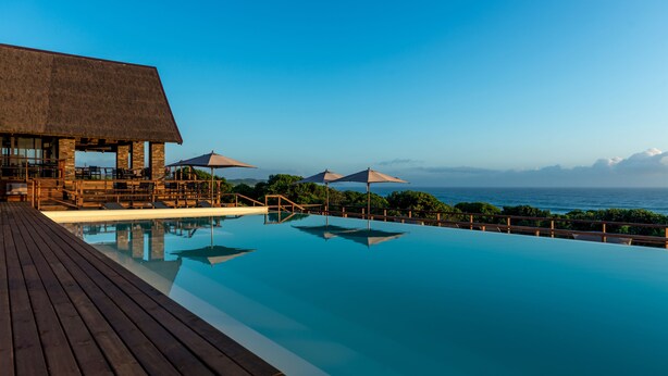 A pool with a wooden deck, umbrellas, and a view of the ocean.