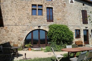 A stone building with a large arched doorway, a small patio with wooden furniture, and a well-maintained garden. - Couette et Tartine (Bozas)