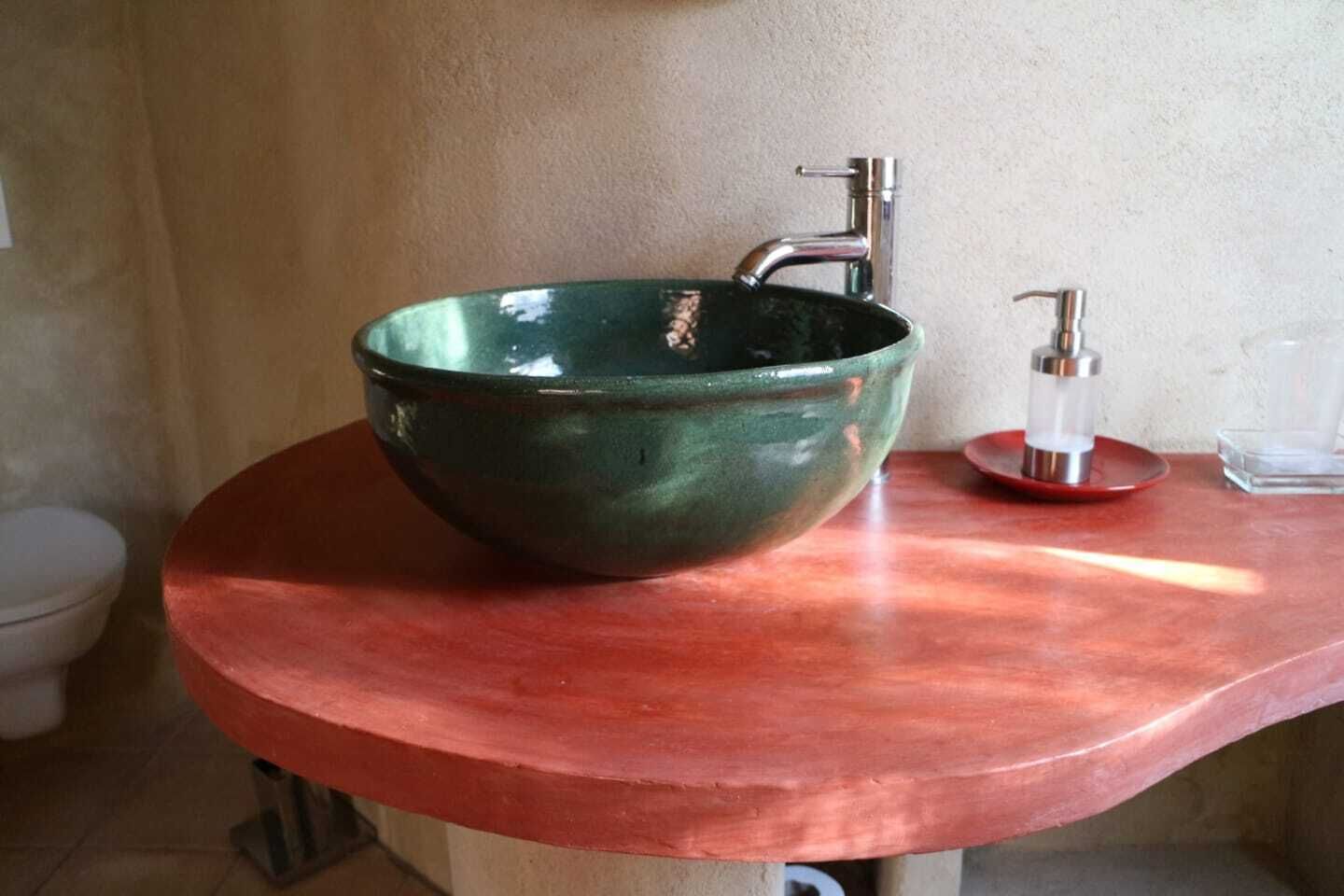 Un évier en céramique verte, doté d’un robinet moderne et d’un distributeur de savon, est installé sur un plan de toilette en cuivre dans une salle de bain.