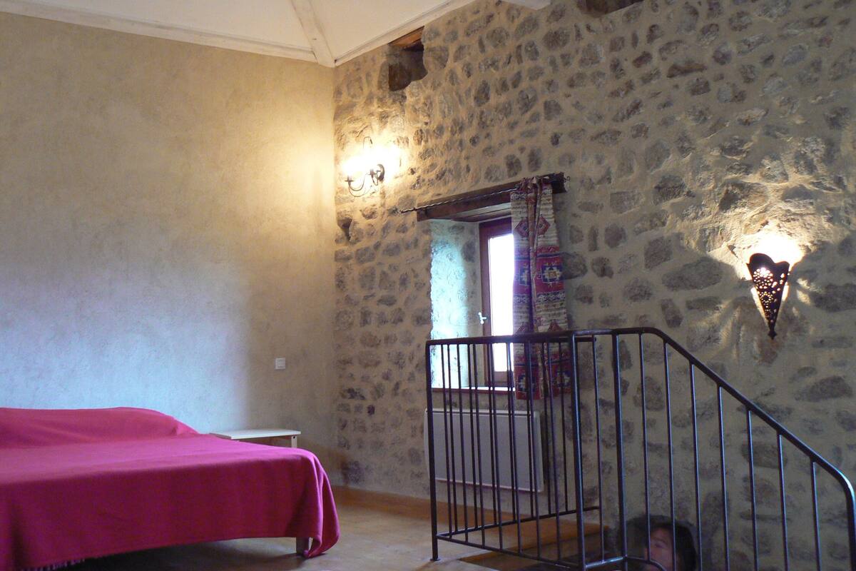 A room with a stone wall, a bed with a pink bedspread, a small table, and a staircase with a railing.