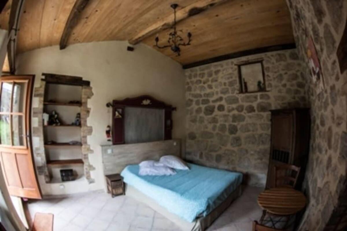 A rustic bedroom with a stone wall, a wooden ceiling, a bed with a blue bedspread, a wooden chair, and a small table.