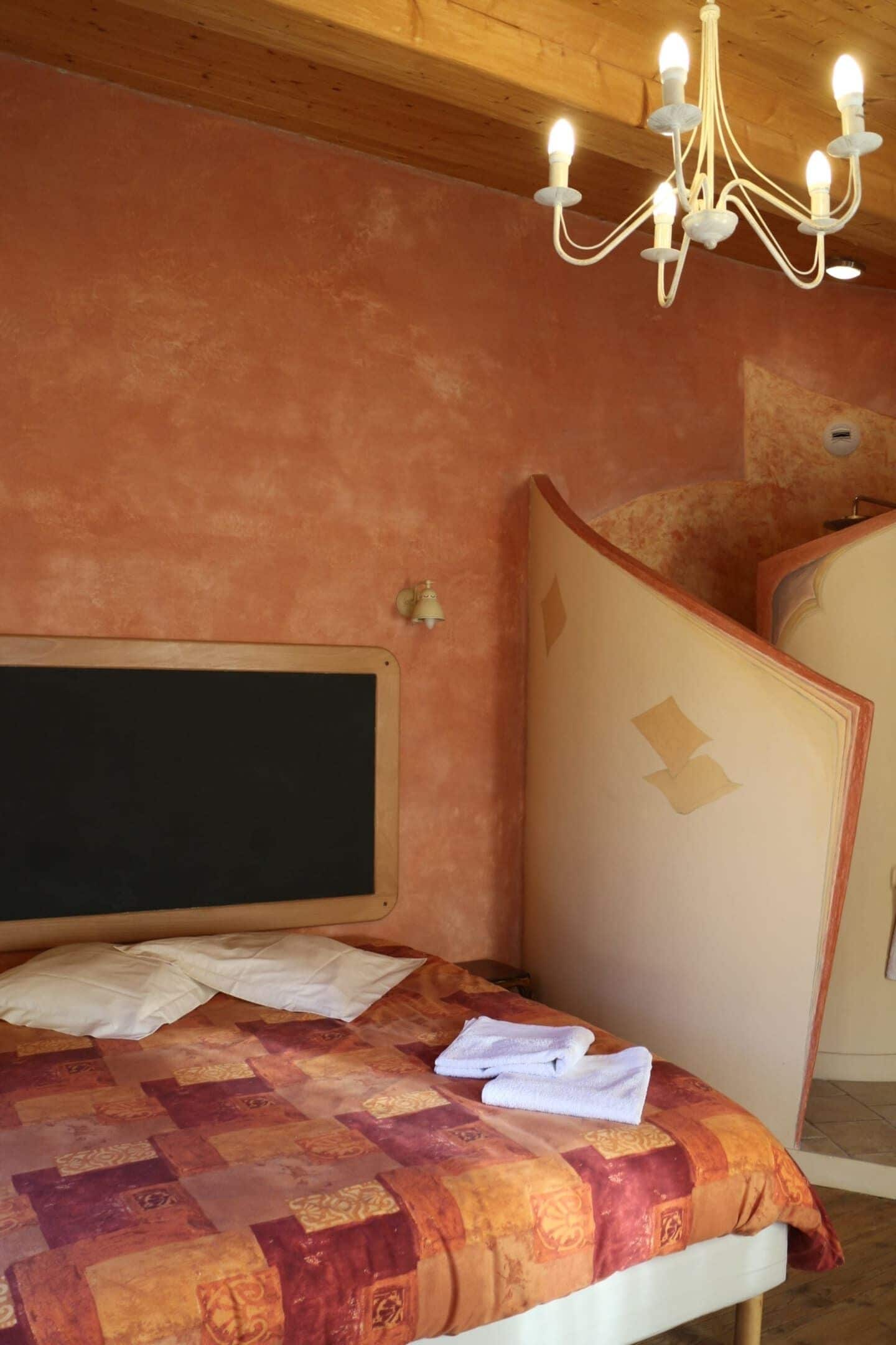 A bedroom with a wooden headboard, a quilted bedspread, a chandelier, and a decorative panel.