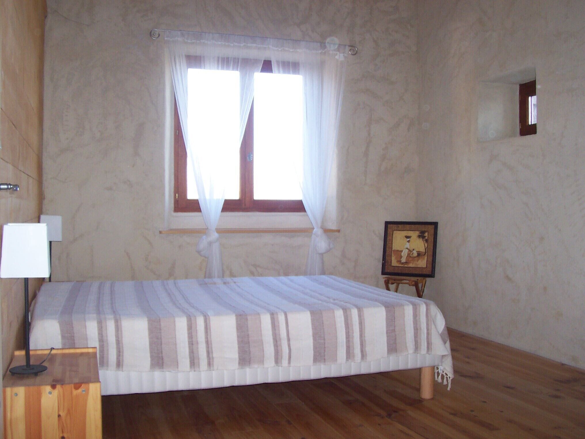 Ein Schlafzimmer mit einem Bett, einem Fenster mit Vorhängen, einer Nachttischlampe und einem kleinen Bildrahmen an der Wand.