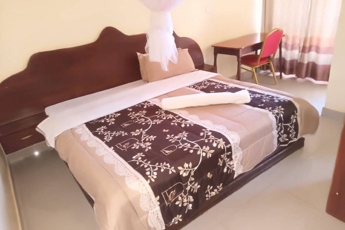 A neatly made bed with a floral bedspread, a bedside table, and a chair in a room with a curtain.