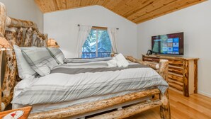 Un dormitorio con techo de madera, cama con ropa de cama a rayas, televisor sobre un mueble de madera y una ventana con cortinas.