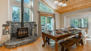 Un comedor con chimenea de piedra, mesa de madera, bancos y ventanas amplias con vistas al exterior.