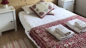 A bedroom with a wooden wall, a bed with white bedding, a red and white patterned throw blanket, two bedside lamps, and a red floral curtain. - Spacious 2-bed apartment in Morzine on ski bus route (Morzine)