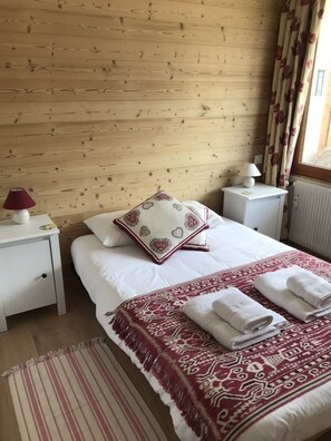Ein Schlafzimmer mit einer Holzwand, einem Bett mit weißer Bettwäsche, einer rot-weißen Decke mit Muster und zwei Nachttischlampen. Ein roter, blumengemusterter Vorhang ist ebenfalls zu sehen.